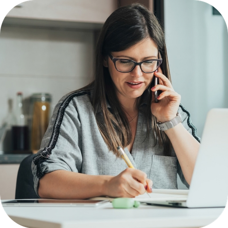 Woman taking notes during a phone call at her kitchen table with a laptop open while learning about weight loss medication